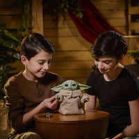 Two children sitting at a table with the Hasbro Star Wars The Mandalorian The Child Animatronic Edition figure.