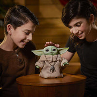 Two children smiling at a Hasbro Star Wars The Mandalorian Animatronic Child figure wearing a silver pendant.