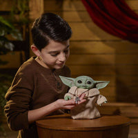 A boy holds the Hasbro Star Wars The Mandalorian Animatronic Child figure, featuring green skin and a tan robe.
