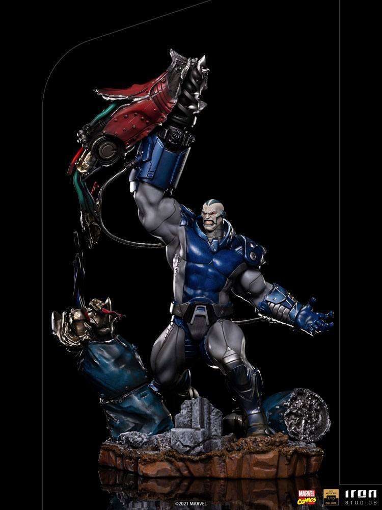 Iron Studios 1/10 Apocalypse Deluxe statue in blue and grey armor, holding a robotic arm aloft on a rocky base.