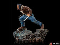 Side view of Iron Studios 1/10 Logan statue showing Wolverine with claws out, wearing a brown jacket on a rocky base.