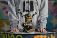 Iron Studios 1/10 scale Leonardo statue standing on a sewer pipe base, held by a person in a grey hoodie.