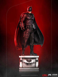 Iron Studios 1/10 The Batman statue in a black tactical suit and cape, standing on a detailed base against a red background.
