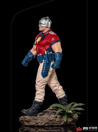 Side view of Iron Studios 1/10 Peacemaker statue wearing a red and blue uniform with a chrome helmet on a rocky base.